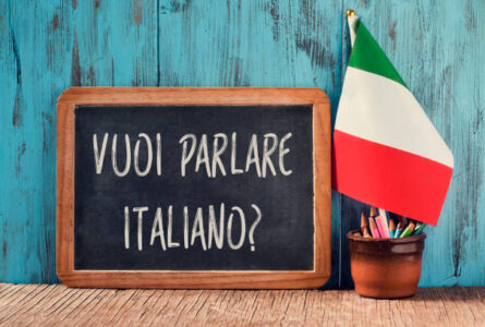 a chalkboard with the question vuoi parlare italiano?, do you want to speak Italian? written in Italian, a pot with pencils, some books and the flag of Italy on a wooden desk